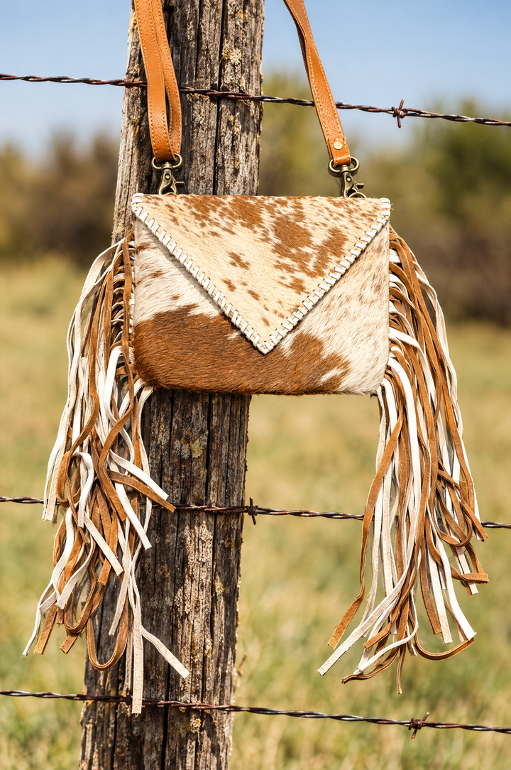 Cowhide fringe crossbody purse hanging on ranch fence